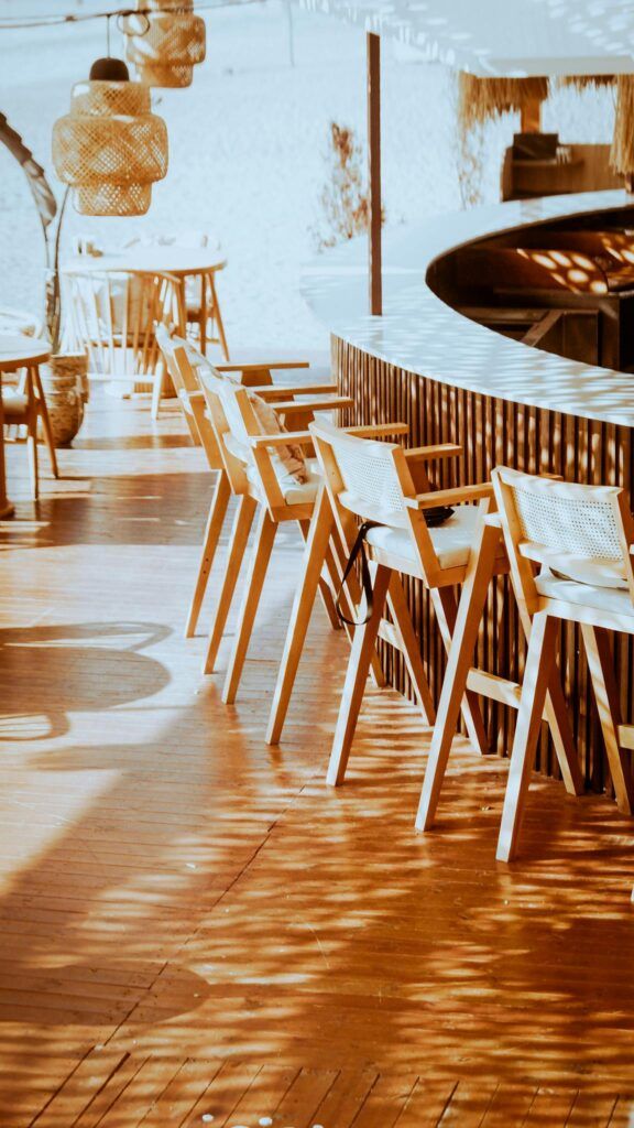 Warm outdoor cafe seating area with wooden chairs and bar under a sunlit canopy.